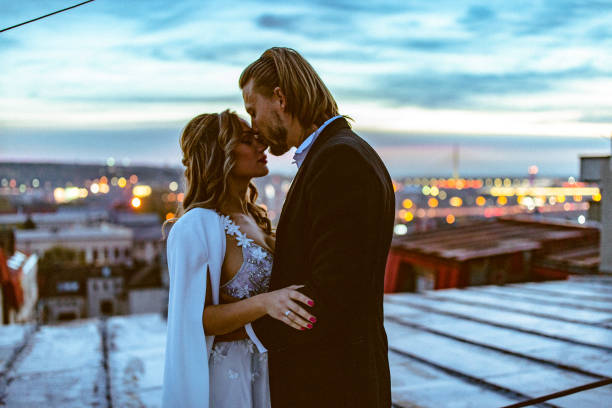 Affectionate bride and groom, embraces and kisses on the rooftop at sunset.