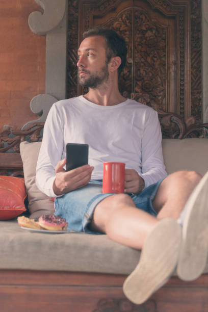 istock Man holding cellphone, coffe mug with donuts on the sofa. 897613622