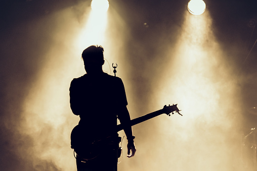 Rock band performs on stage. Guitarist plays solo. silhouette of guitar player in action on stage in front of concert crowd. Smoke. Light