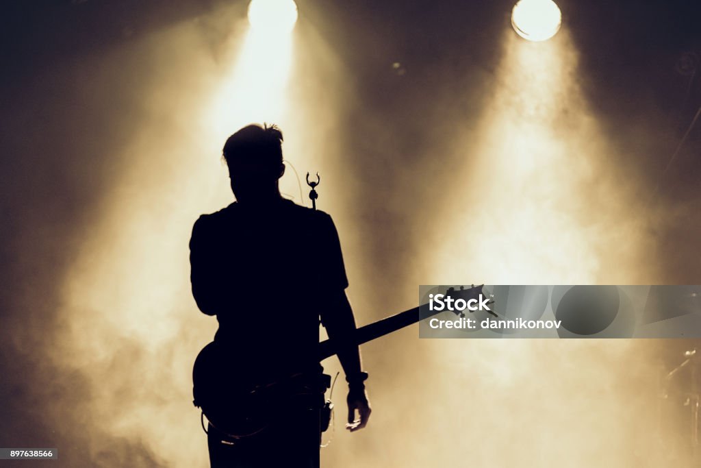Rock band performs on stage. Guitarist plays solo. silhouette of guitar player in action on stage behind lights. Rock band performs on stage. Guitarist plays solo. silhouette of guitar player in action on stage in front of concert crowd. Smoke. Light Musician Stock Photo