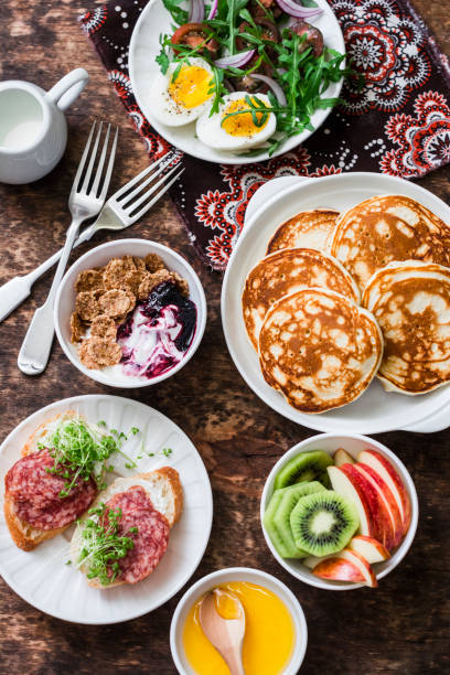 istock Breakfast table - greek yogurt with whole grain cereals and berry sauce, pancakes, arugula, cherry tomatoes, boiled eggs salad, kiwi, apples fruit, salami and cream cheese sandwiches on a wooden background, top view. Flat lay 897642716