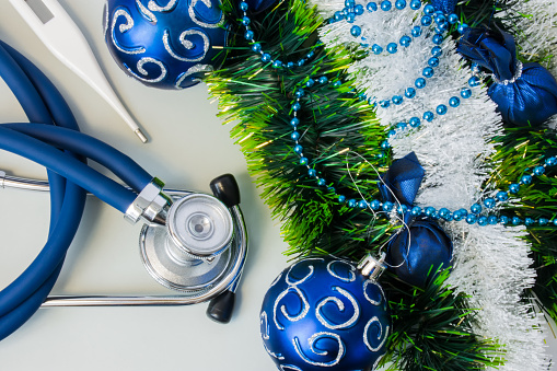 Christmas and New Year decorations near medical equipment. Medical stethoscope and thermometer lying near artificial snow with glitter, toys and blue balls on Christmas tree. New Year in Medicine