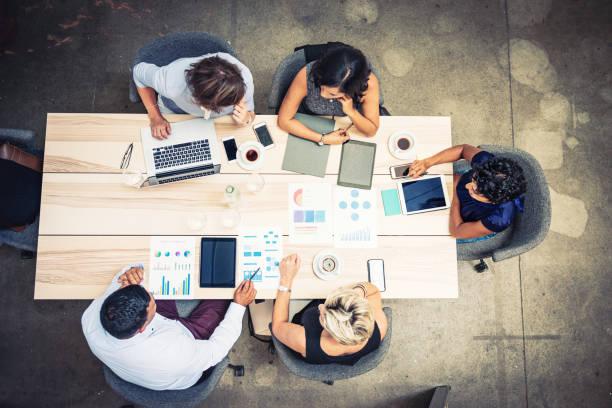 A group of five people, seated around a light wooden table, are engaged in a collaborative discussion, surrounded by laptops, tablets, smartphones, and various documents featuring charts and graphs, with coffee cups also present on the table.