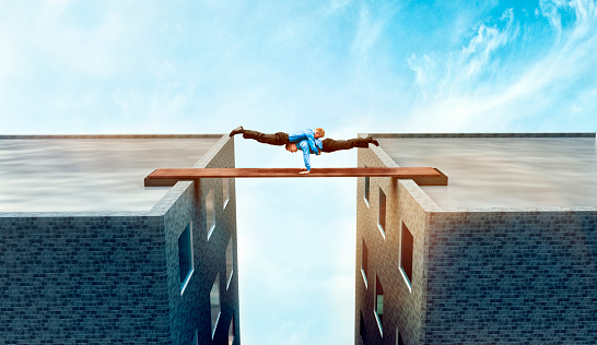 Two acrobatic businessmen support each other while keeping the balance on a plank between two buildings. Concept of teamwork and trust in each other while the stakes are high.