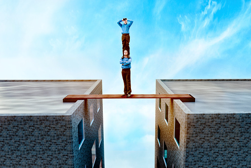 Two acrobatic businessmen support each other in the attempt to find new opportunities. One man stands on top of the other and holds binoculars. The men stand on a small wood board between two office buildings.