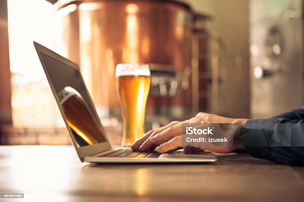 Brewer using laptop at micro brewery Close up of master brewer hands using laptop computer at micro brewery. Man sitting at table with laptop and beer glass. Beer - Alcohol Stock Photo