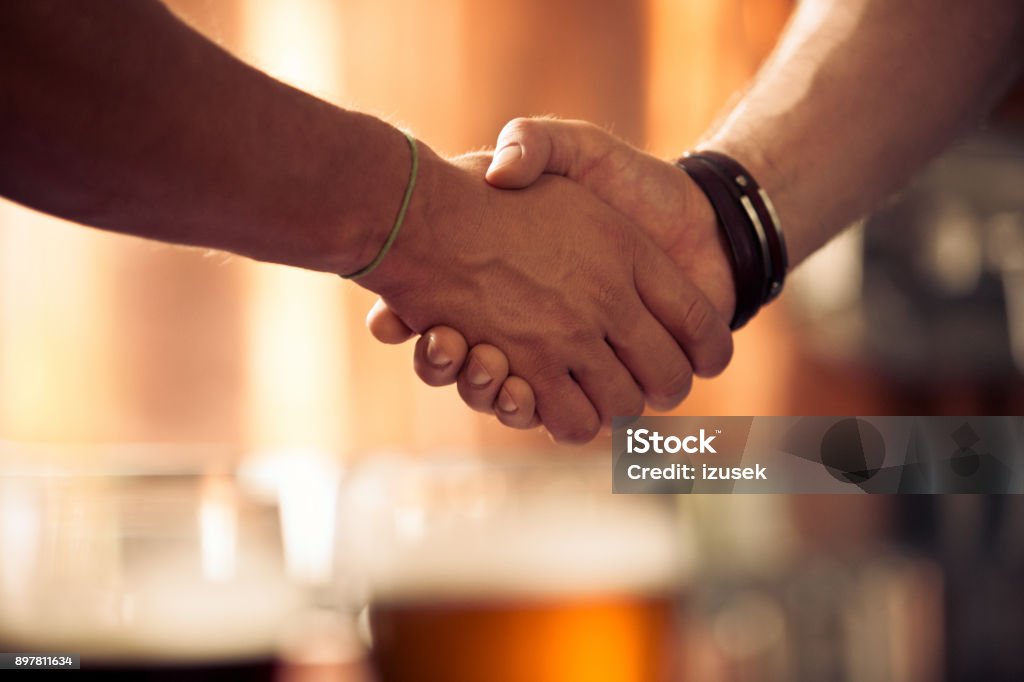 Businessmen hand shake at micro brewery Close up of two businessmen shaking hands at micro brewery. Focus on shaking hands with beer glasses in background. Handshake Stock Photo