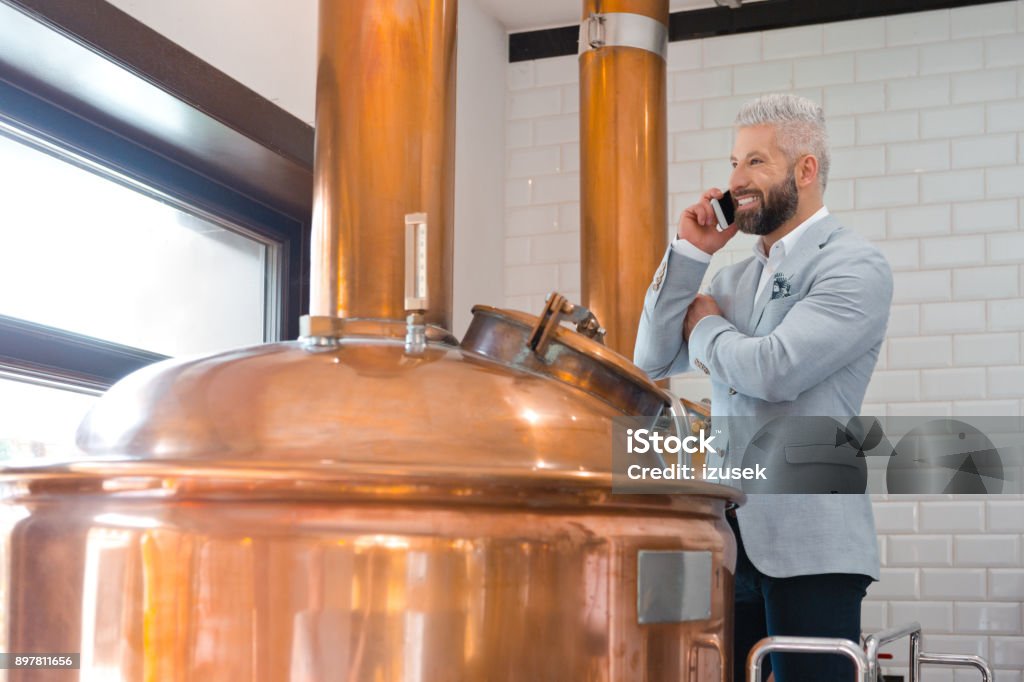 Microbrewery owner using a mobile phone Mature businessman standing by a copper vat in brewery talking on mobile phone. Micro brewery owner using cell phone in fermenting section of beer manufacturing factory. Adult Stock Photo