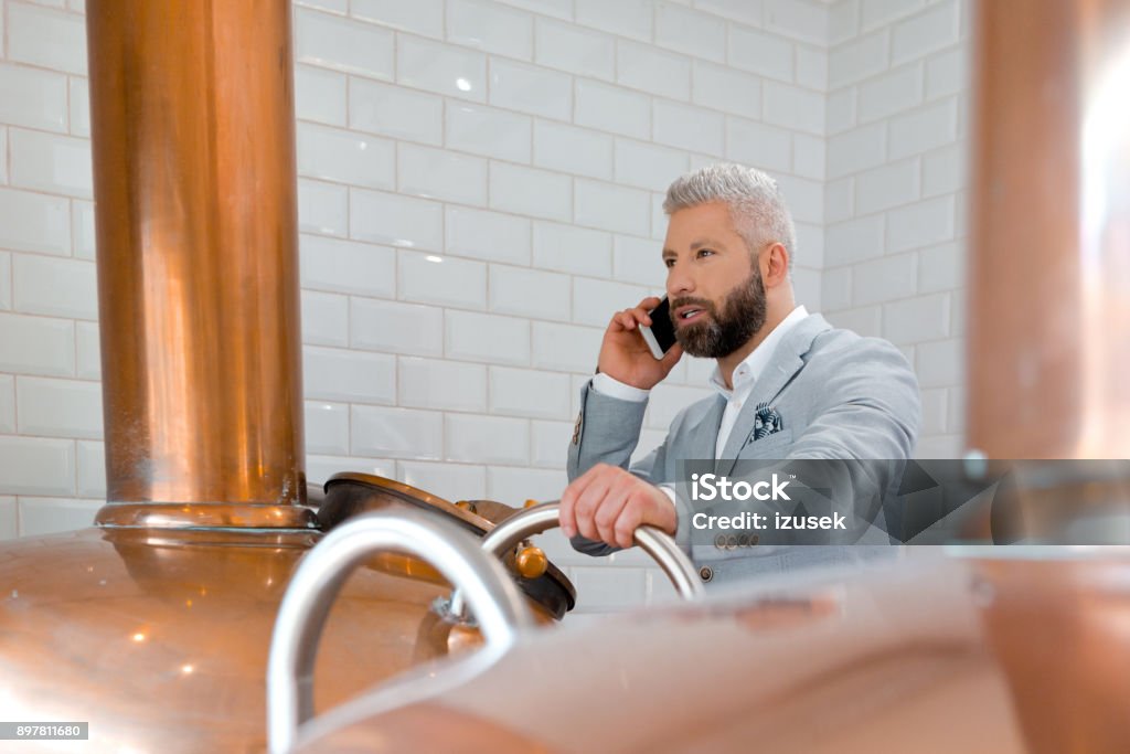 Businessman using mobile phone in his micro brewery Businessman talking on mobile phone while standing by a copper vat in brewery. Micro brewery owner in fermenting section of beer manufacturing factory making a phone call. Adult Stock Photo