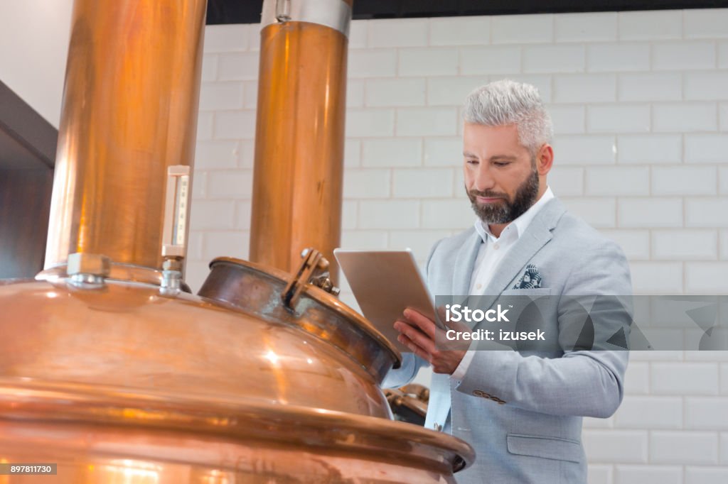 Microbrewery owner using a digital tablet in fermenting section Mature businessman with digital tablet standing by a copper vat in brewery. Micro brewery owner with a digital tablet in fermenting section of beer manufacturing factory. Fermenting Stock Photo