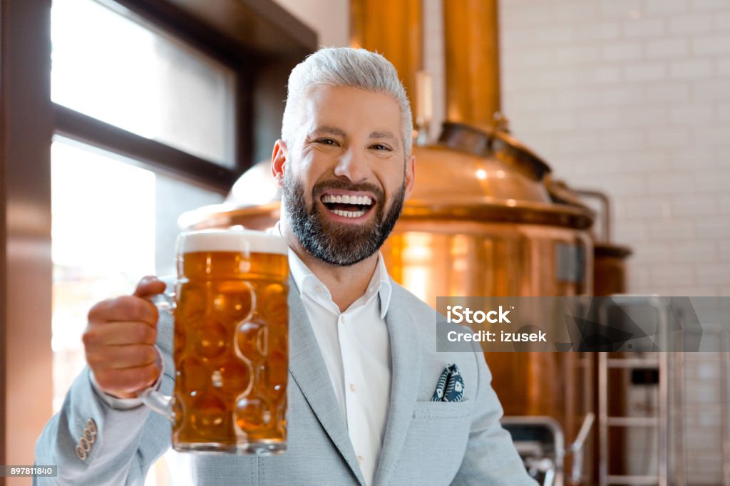 Laughing businessman holding a beer mug in microbrewery Portrait of laughing mature businessman holding a beer mug in microbrewery. Elegant man in suit with beer glass looking at camera and laughing. Adult Stock Photo