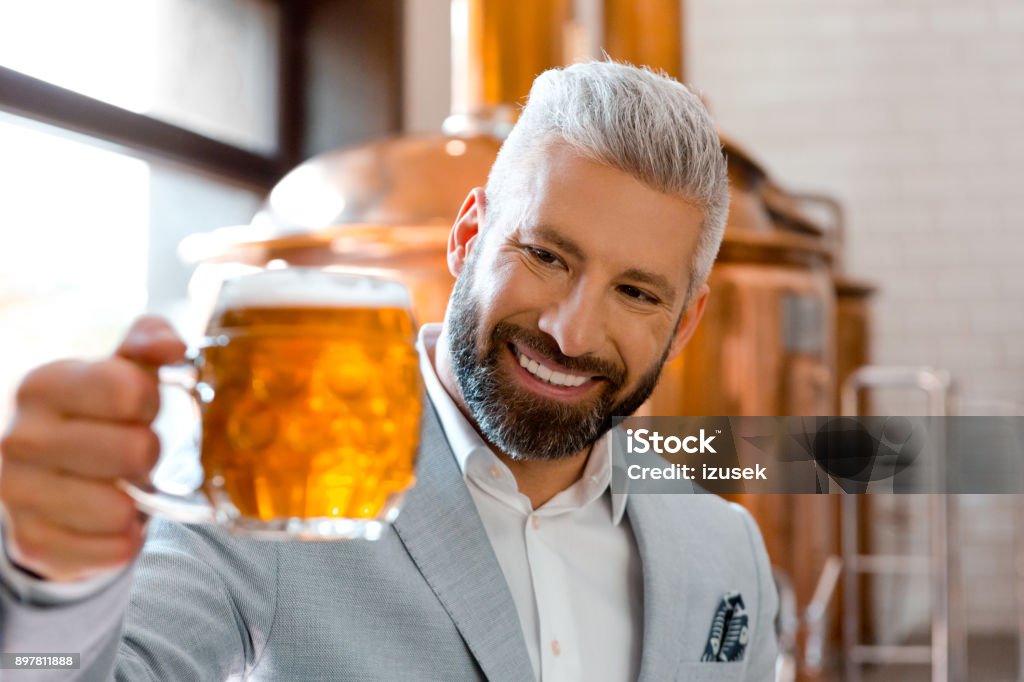 Smiling microbrewery owner holding a beer mug in his pub Smiling microbrewery owner holding a beer mug in his pub. Businessman looking at fresh beer glass at his brewery. Adult Stock Photo