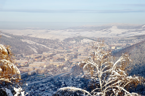 Winter snow mountain valley with Gorno-Altaisk town and frozen rime trees in the foreground with ranges of snow hills on the background under blue sky Altai Mountains Siberia, Russia