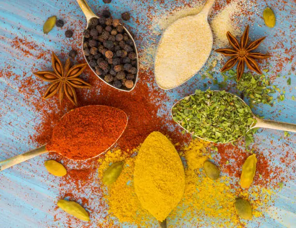 Photo of Milled spices - garlic, turmeric, paprika, anise, oregano, cardamom.  Round of golden spoons on  blue wooden table. Top view, close-up