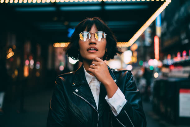 joven inconformista vestido moda en gafas con reflejo de luz de la ciudad de noche fascinado con hermosa iluminación, hermosa mujer en chaqueta de cuero preguntando mientras está parado sobre fondo urbano en la noche - city urban scene night lighting equipment fotografías e imágenes de stock