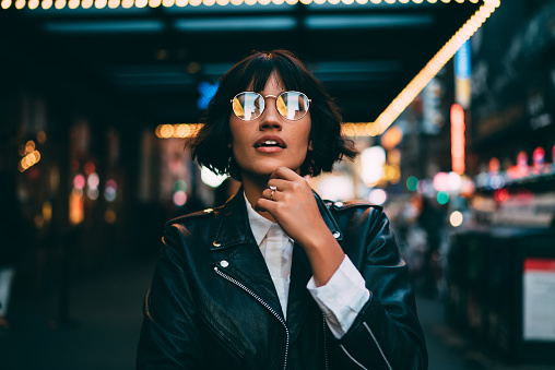 Young trendy dressed hipster in eyewear with night city light reflection fascinated with beautiful illumination, gorgeous woman in leather jacket wondering while standing on urban background at evening