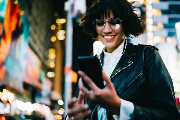 istock Cheerful hipster girl in eyewear using mobile application for navigating in night megalopolis listening music in earphones,happy young woman reading SMS spending time in evening city with illumination 897963126