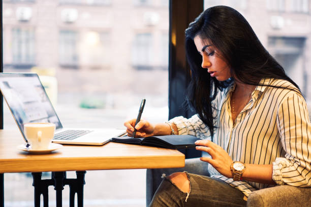 Attractive contemplative businesswoman pondering about creative ideas for startup project while making some notations to black notepad.Intelligent female student sitting in cafeteria and studying