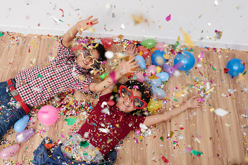 Cheerful kids lying on the floor in funny glasses, sparkles and balloons everywhere