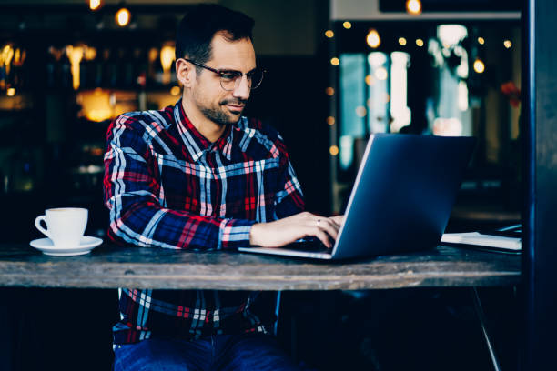istock Concentrated young man copywriter in stylish eyeglasses keyboarding information on modern laptop computer sitting in coffee shop interior.Male graphic designer working freelance at digital netbook 897979554