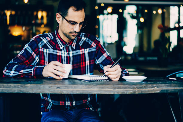 istock Young hipster guy in stylish eyeglasses writing down checklist in notepad during coffee break.Student in spectacles noting text information in notebook sitting with cup of tasty beverage in cafe 897979574