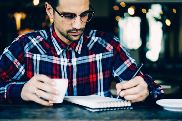 istock Cropped image creative young man in stylish eyewear organizing plans and writing down in notebook sitting in cafeteria interior with cup of beverage.Male student in spectacles noting text in notepad 897979714