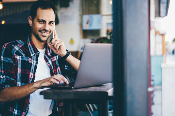 istock Half length portrait of happy young man talking in roaming via mobile tariffs on modern smartphone device while sitting indoors in cafe with laptop computer and working remotely on new project 897979730