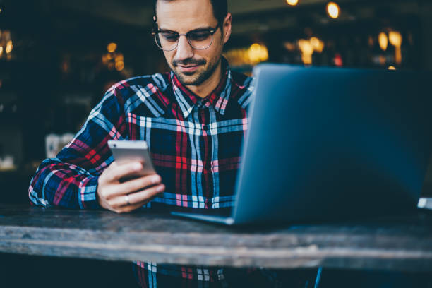 istock Cropped image of bearded student in optical spectacles reading blog on telephone device while preparing for seminar sitting at laptop computer for searching new information for productive work 897979748