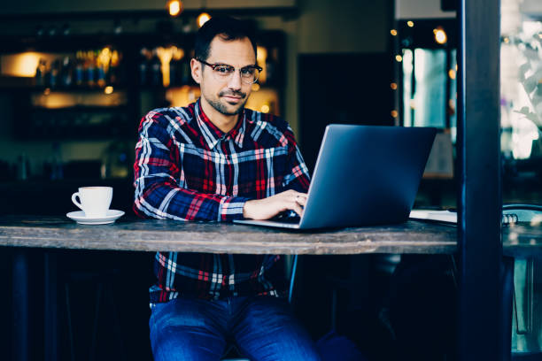 istock Portrait of young bearded businessman in optical eyeglasses looking at camera while working remotely on modern digital laptop computer.Handsome male typing text on keyboard of netbook sitting in cafe 897979764