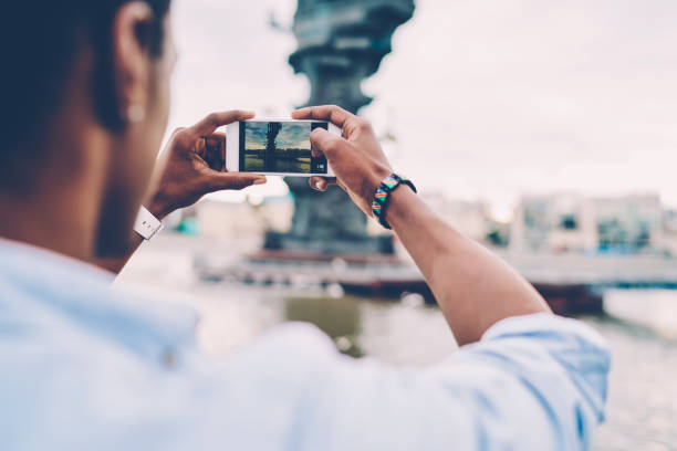 istock Selective focus on man's hands holding modern cellphone and trying to make good picture of showplace near to river.Cropped back view image of tourist making photo on cellular during strolling outdoors 897981426