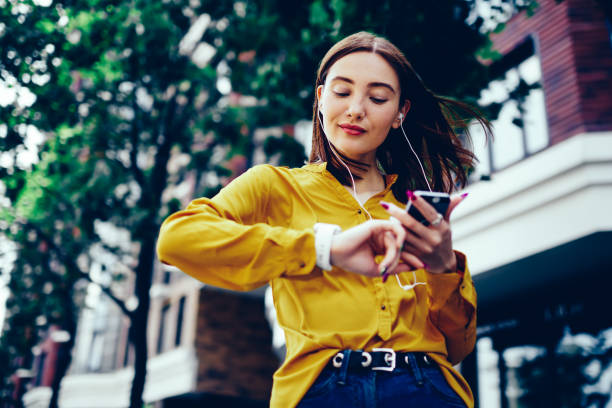 istock Charming redhead female with windy hair checking time at wristwatch while strolling at summer street.Young cute hipster girl dressed in casual clothing listening to favorite playlist in website 897987884