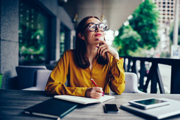 Pondering cute talented student in cool spectacles looking away and thinking while preparing for upcoming exams.Pensive charming redhead hipster girl writing down information to notepad in cafe