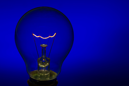Glass light bulb with burning filament upright with bright blue background