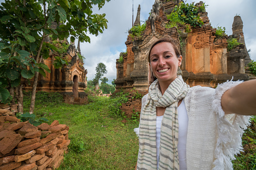 Caucasian young woman takes selfie with ancient temples pagodas and monastery coming out from the lush greenery. People travel concept.