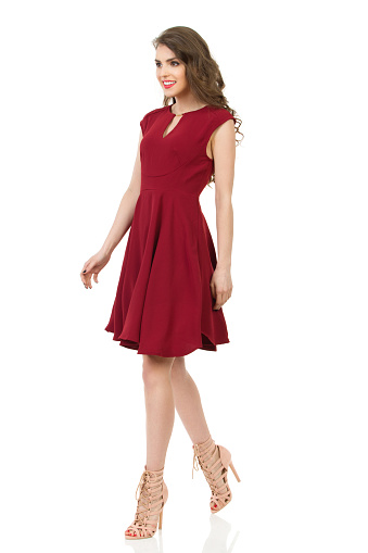 Young woman in red dress and high heels is walking, looking away and smiling. Front side view. Full length studio shot isolated on white.