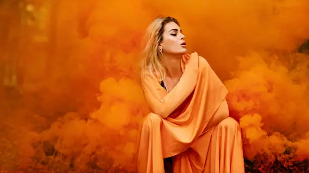 Photo of Beautiful young blond female woman dancing and posing in orange smoke in outdoor countryside setting