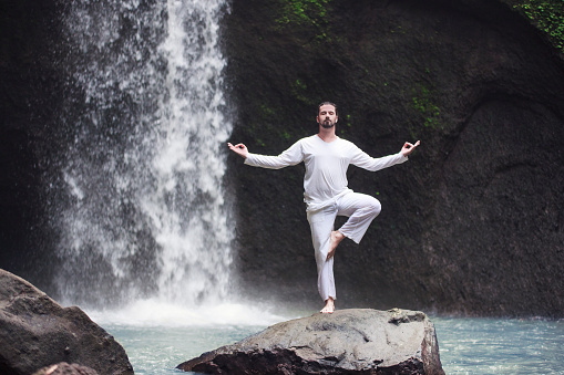 Man standing in meditation yoga on rock at waterfall in tropical Bali
