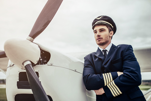 Young handsome man pilot in uniform is standing near small private plane.