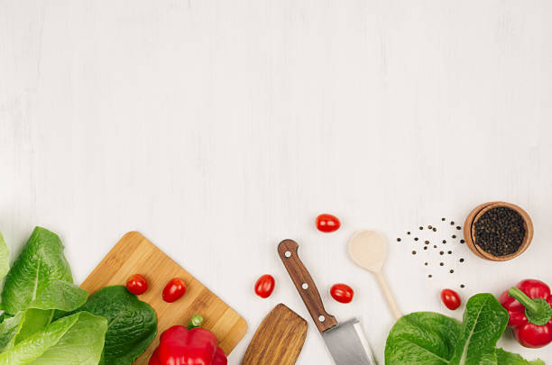 Fresh green vegetables, greens, red cherry tomatoes and kitchenware on soft white wood board, border, top view.