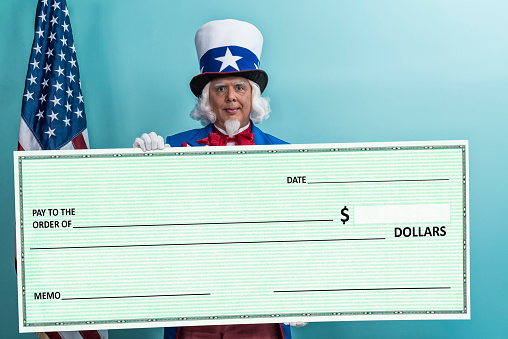 A patriotic Uncle Sam character holding a large green blank check, looking at the camera. He is wearing a red, white and blue coat and top hat that has a blue ban and white star while standing in from of a teal wall with an American flag in the background. Concept - Tax cuts/tax refunds