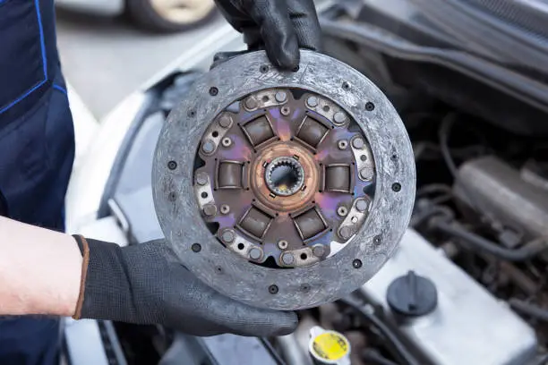 Auto mechanic wearing protective work gloves holds old clutch disc over a car engine