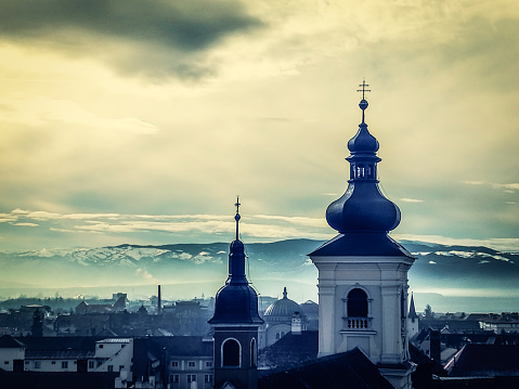 Horizontal color image depicting the ancient, medieval skyline of Sibiu, a city in the Transylvania region of Romania. In the foreground we can see two orthodox church spires, while beyond we can see traditional Germanic architecture of the town's buildings and houses spread out. In the distance the snow-capped Carpathian mountains provide a beautiful backdrop on this winter Decemver day. Room for copy space.