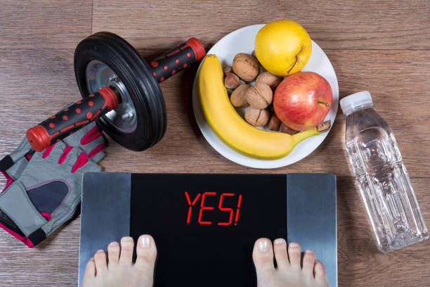 Female feet on digital scales with word yes surrounded by sport accessories (AB roller wheel, fitness gloves), plate with healthy food and water bottle. Concept of active healthy lifestyle. stock photo