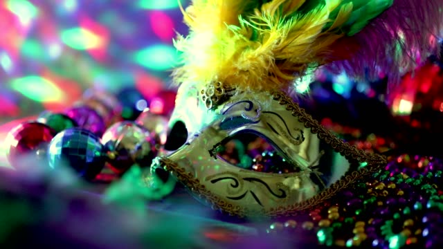 Mardi Gras or Rio Carnival mask and colorful carnival decorations.  Scene includes: gold feathered and sequined mask, colored party lights, and beads.  Objects lie on wooden table.  No people.