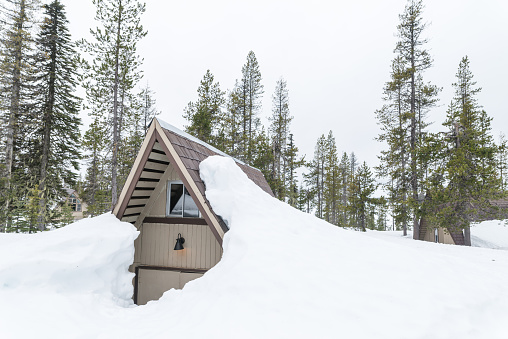 A vintage A-frame cabin is half buried in snow drifts, aside from the driveway that has been cleared. The peak is poking out from the top and the top windows are visible. The area is surrounded by snow drifts and pine trees.
