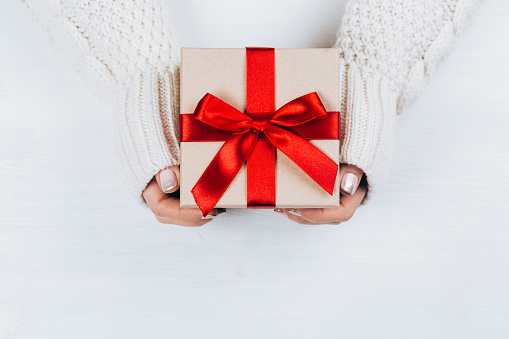 Female hands holding present with red bow on white rustic background. Festive backdrop for holidays: Birthday, Valentines day, Christmas, New Year. Flat lay style