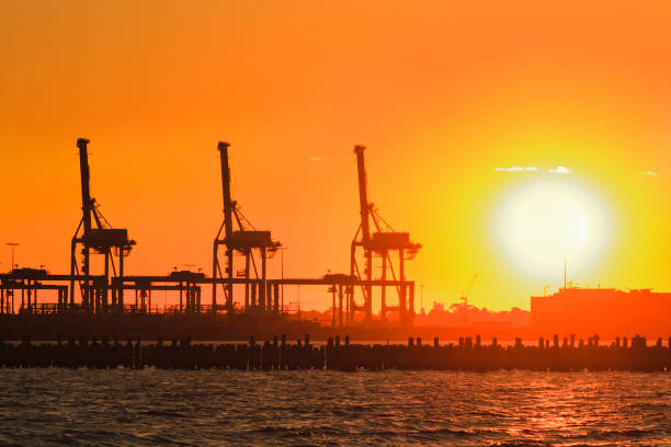 Orange setting sun in line with tall industrial cargo cranes in Melbourne Port dock terminal against distant horizon.