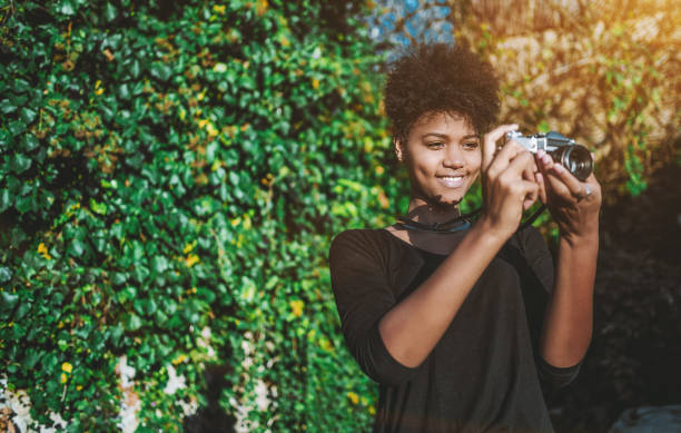 Black girl using vintage film cam in the garden Happy lovely African American female hipster photographer is taking picture of cityscape while staying in front of hedgerow; sunny day; with copy space zone for logo, your advertising or text message quickset stock pictures, royalty-free photos & images