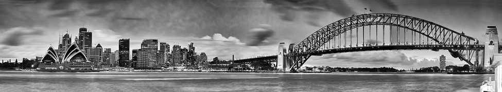 Panoramic view of Sydney cityscape at sunset across Harbour from Kirribilli. CBD landmarks and Harbour bridge are under illumination in black white.