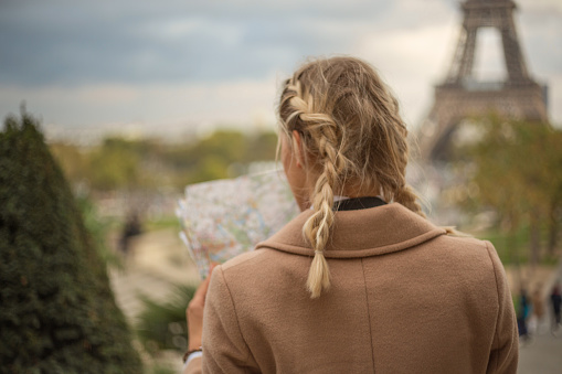 Beautiful blond woman in Paris looking at street map for directions. Tourist looking for sightseeing and location inn the city. Shot at Trocadero, view on the Eiffel Tower, France.\nPeople travel exploring city concept